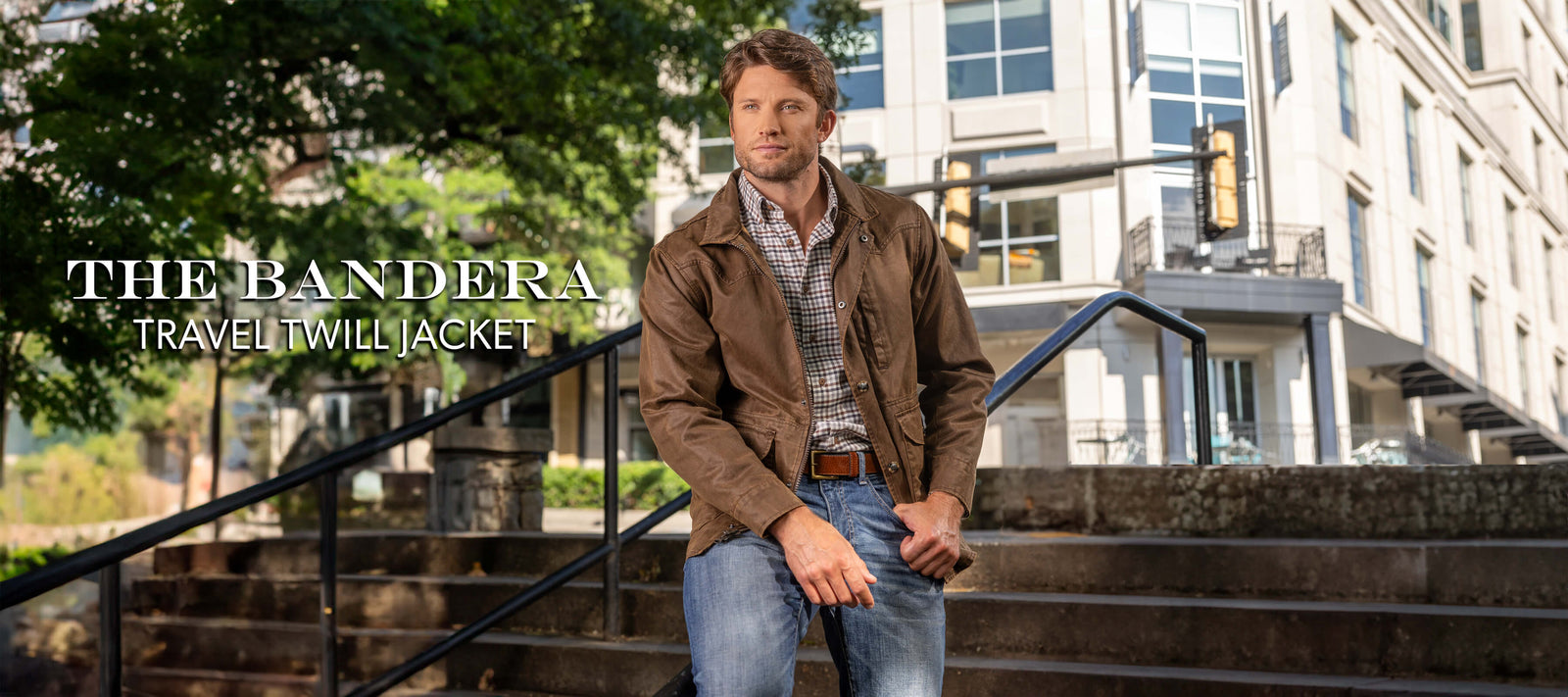 Picture of a tall, handsome man standing on a balcony with black metal railing overlooking a park. He's wearing a brown leather jacket, white shirt and khaki pants.