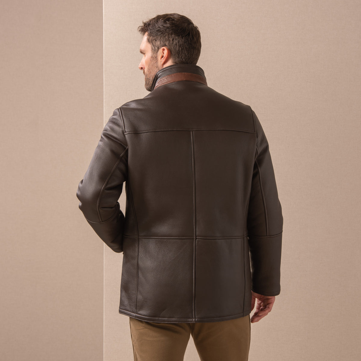 Man wearing a dark chocolate Merino lambskin shearling coat against a beige linen background - back view