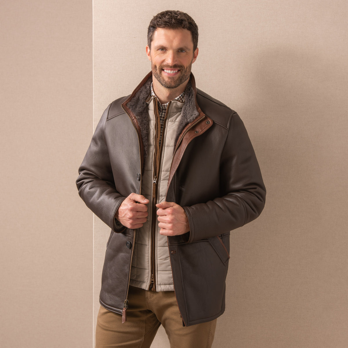 Man wearing a dark chocolate Merino lambskin shearling coat against a beige linen background - front view