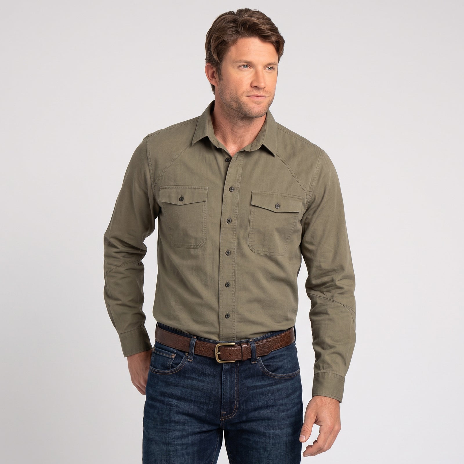 A man wearing an olive green long-sleeve casual button-down shirt with two chest pockets, paired with dark blue jeans and a brown belt, standing against a light gray background.