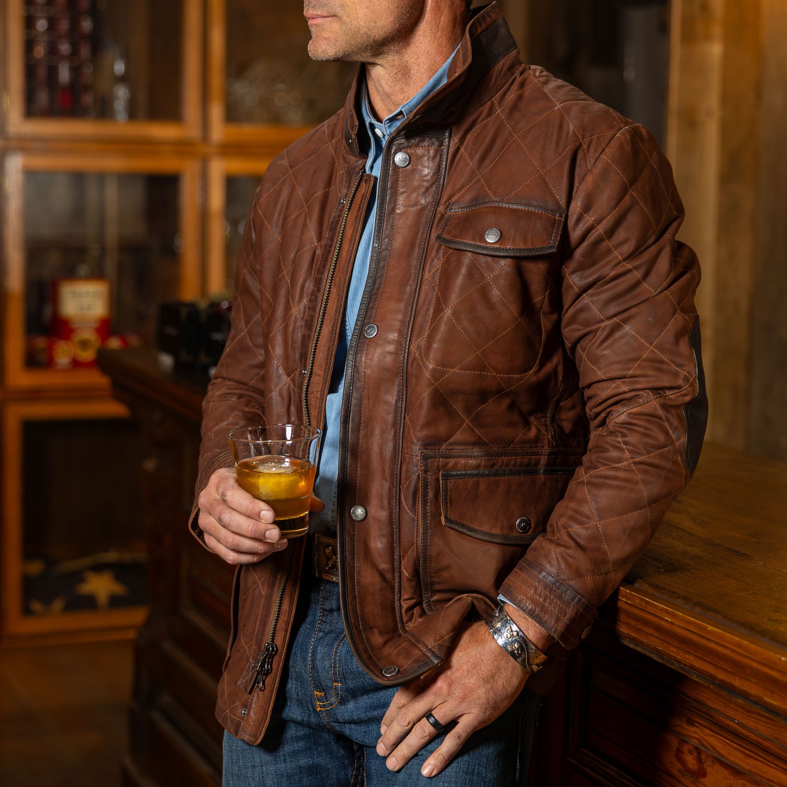 Model wearing the men’s Sedona Waxed Goat Suede Jacket in Bourbon with removable shearling collar, worn unzipped over blue shirt with jeans.