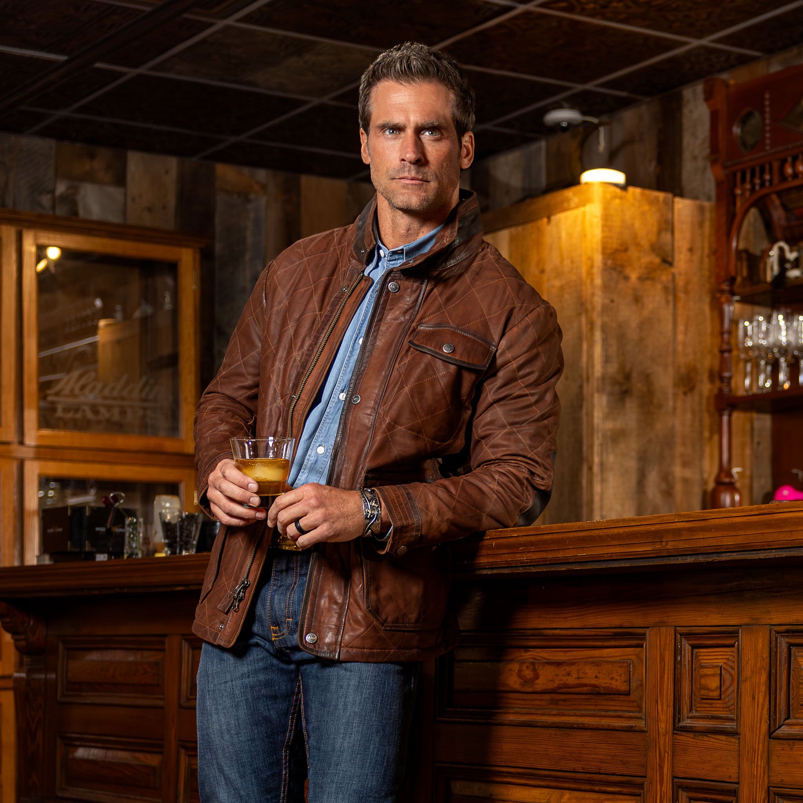 Model wearing the men’s Sedona Waxed Goat Suede Jacket in Bourbon with removable shearling collar, worn unzipped over blue shirt with jeans.