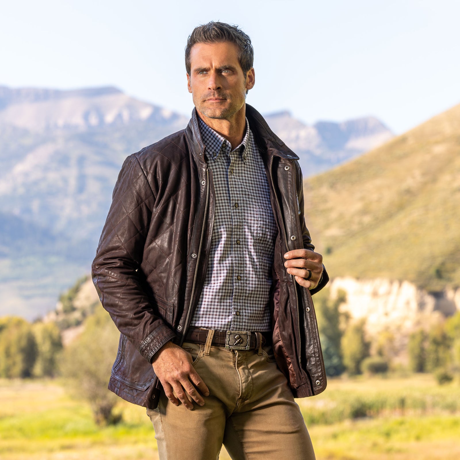 Model wearing the men’s Sedona Waxed Goat Suede Jacket in Chocolate unzipped, standing in a mountain landscape with check shirt and khaki pants.